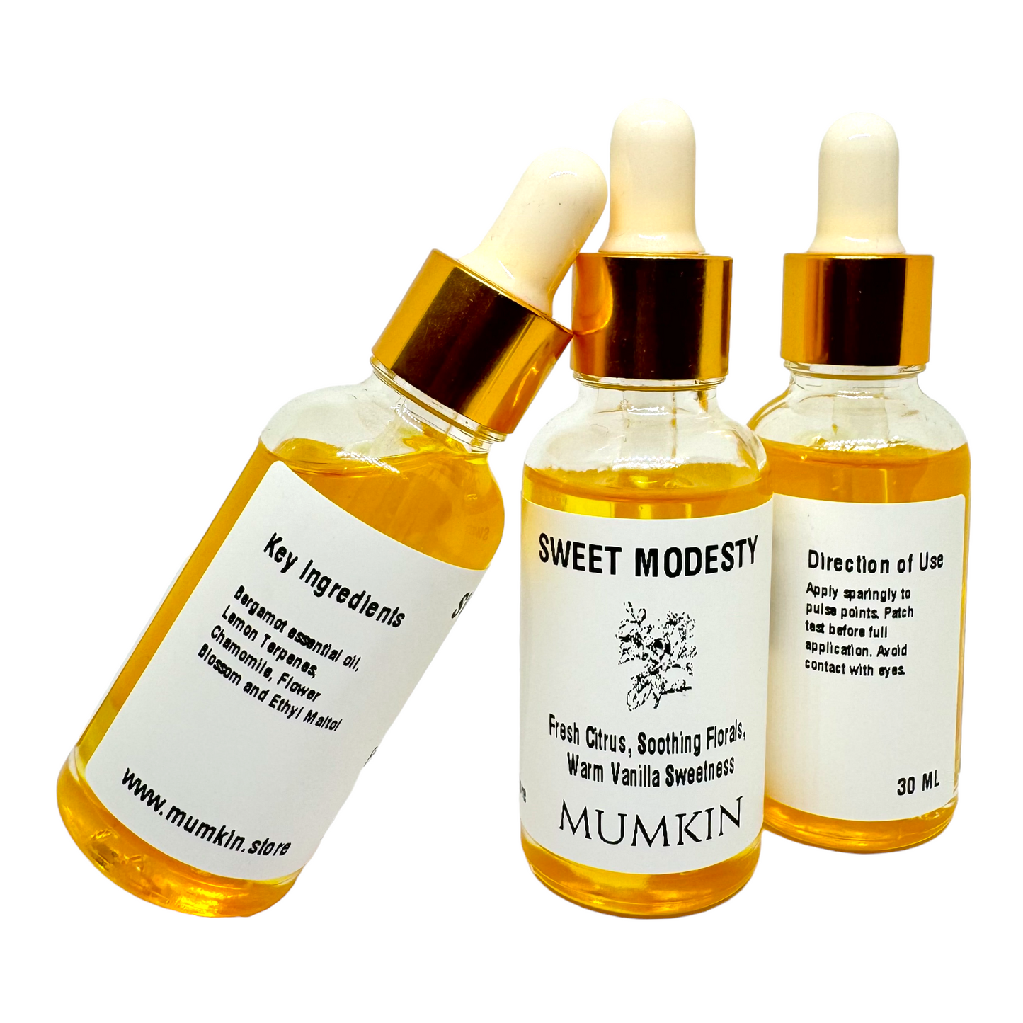 Three bottles of "Sweet Modesty" fragrance oil by Mumkin, each showing different label details: the main scent notes, key ingredients, and usage directions. The bottles contain golden-hued oil and have gold-capped droppers with white rubber tips against a plain white background.