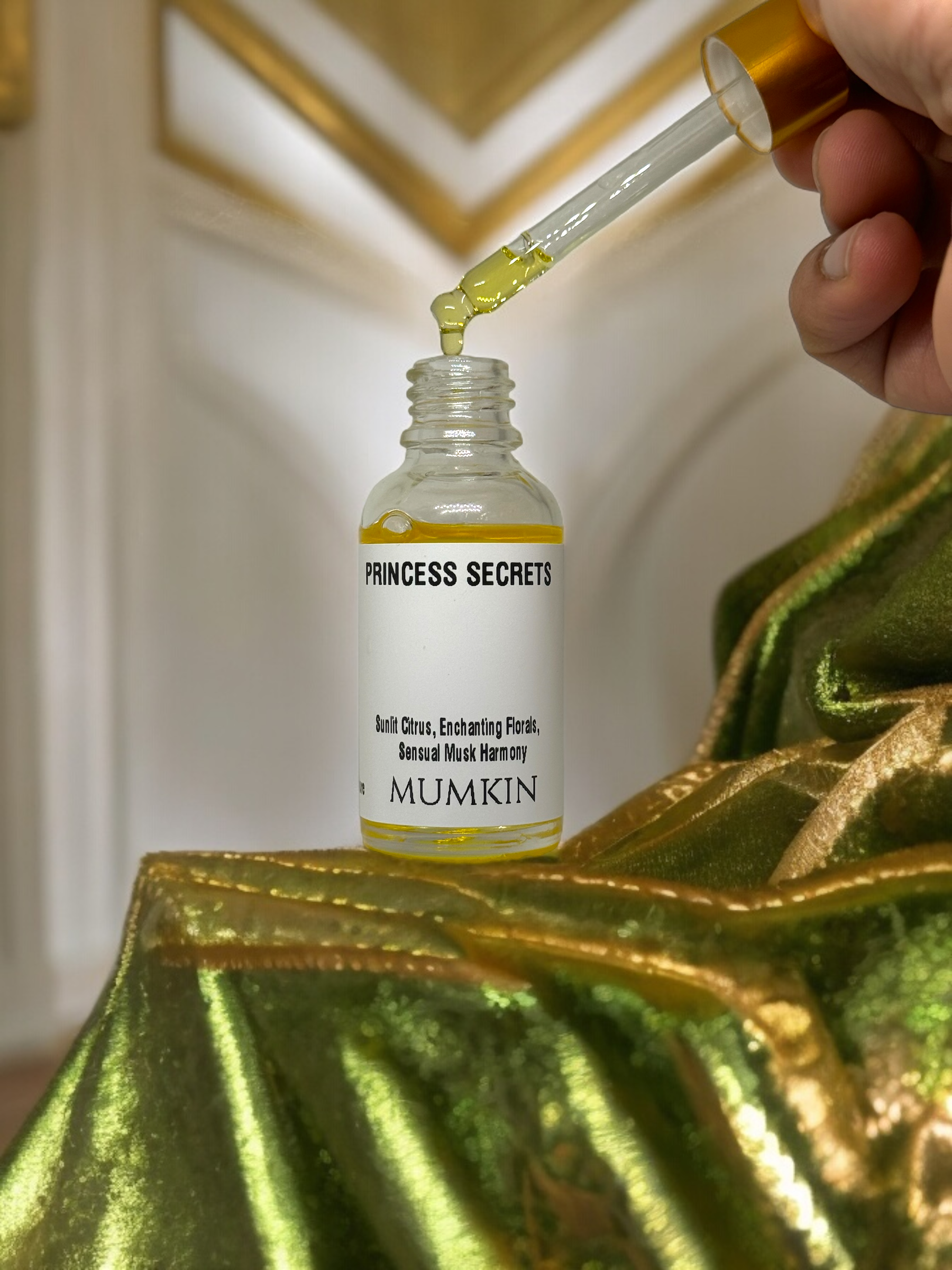 This is a beautiful and luxurious product shot of the "PRINCESS SECRETS" fragrance oil by MUMKIN. The elegant gold and white packaging contrasts well with the green and gold fabric in the background, creating a sophisticated and high-end aesthetic. The addition of the dropper and hand adds a dynamic element, emphasizing the product's application and texture.
