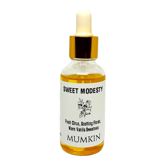 A 30ml glass dropper bottle labeled 'Sweet Modesty Fragrance Oil ' by Mumkin, featuring a gold cap and white label with black text. The fragrance notes include Fresh Citrus, Soothing Florals, and Warm Vanilla Sweetness. The liquid inside the bottle is golden yellow.