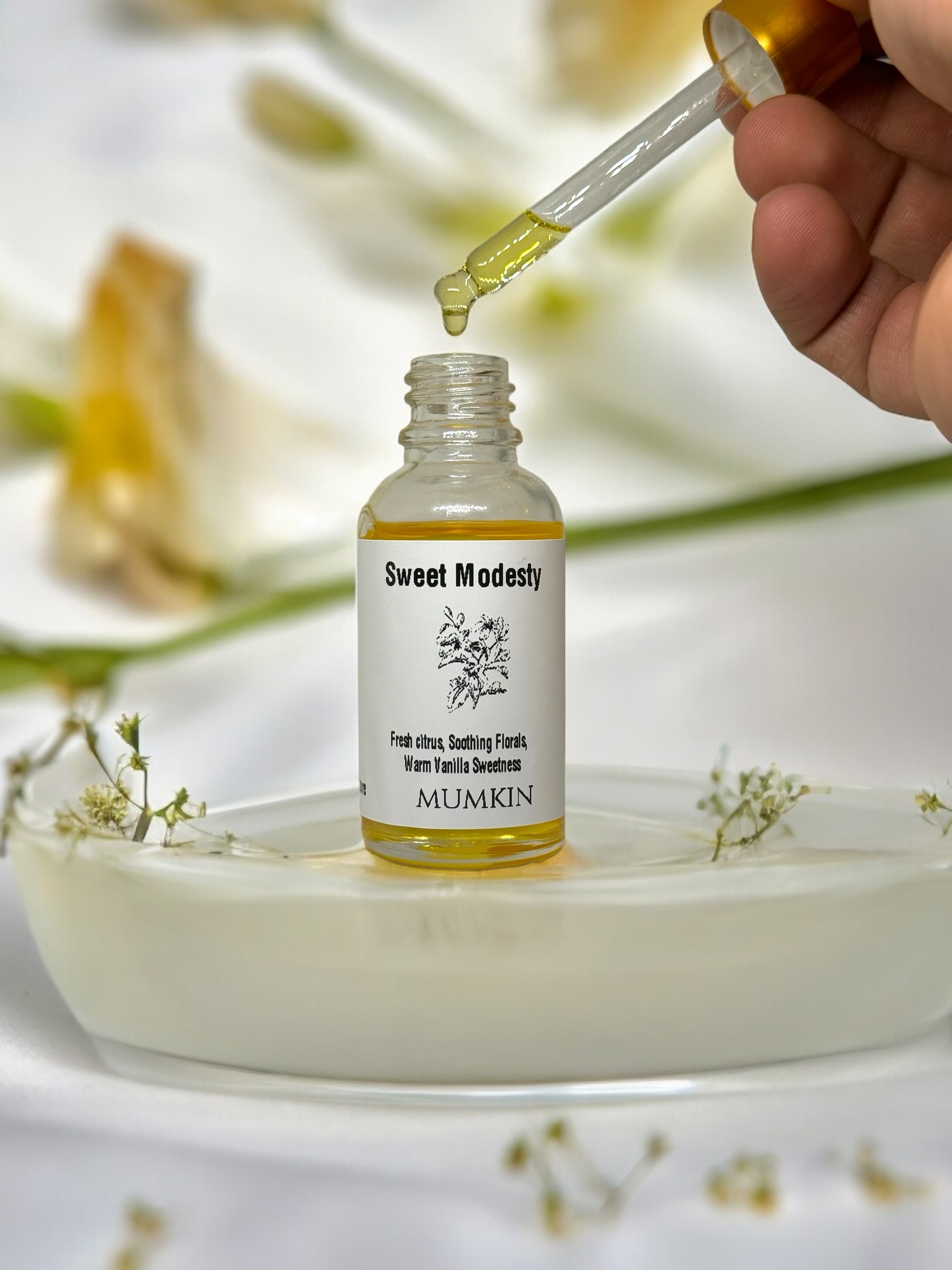 "A close-up of a dropper bottle labeled 'Sweet Modesty' by MUMKIN, with golden-hued oil being dispensed from a glass dropper. The label describes the fragrance as a blend of fresh citrus, soothing florals, and warm vanilla sweetness. The bottle is placed on a white ceramic dish, surrounded by delicate white flowers, with a soft-focus background.